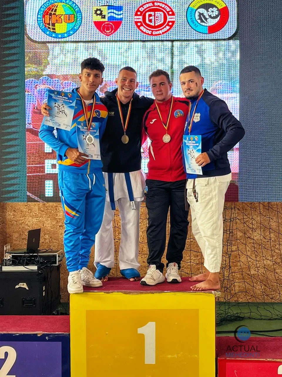 Patru bărbați stau pe un podium, purtând costume de karate și medalii, în fața unui ecran mare cu diverse embleme colorate.