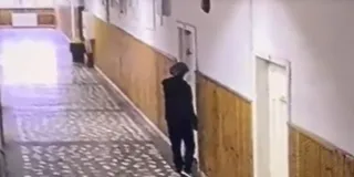 O persoană îmbrăcată în negru merge pe un coridor cu pereți din panouri de lemn și podea cu modele geometrice.