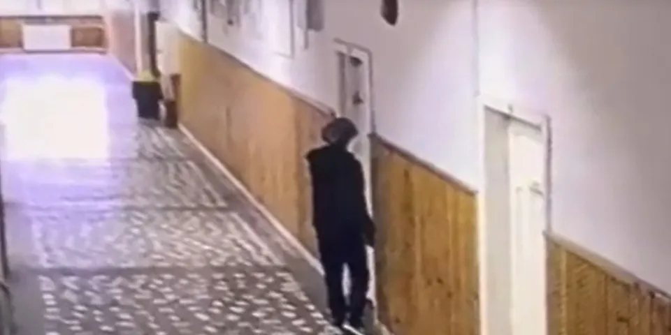O persoană îmbrăcată în negru merge pe un coridor cu pereți din panouri de lemn și podea cu modele geometrice.