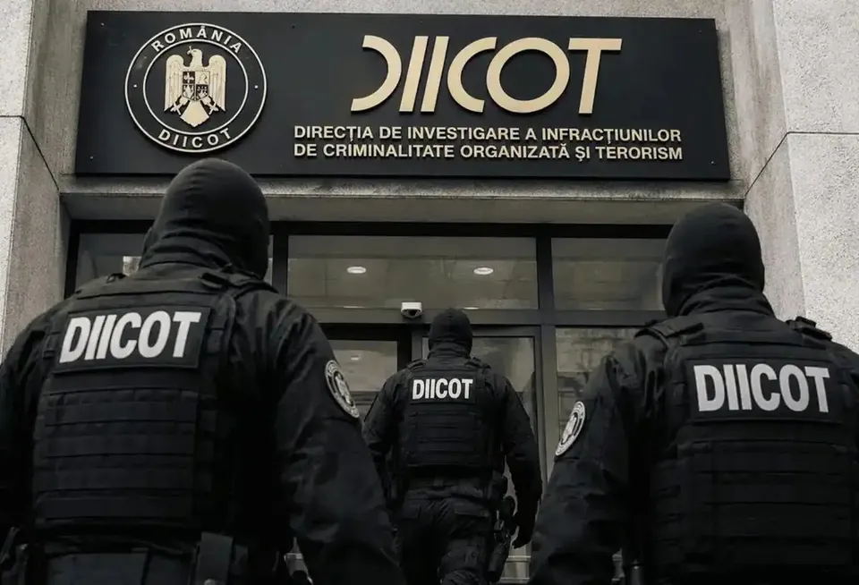 Trei polițiști în uniforme negri cu inscripția DIICOT pe spate stau cu spatele în fața unei clădiri cu un panou negru pe care scrie DIICOT.