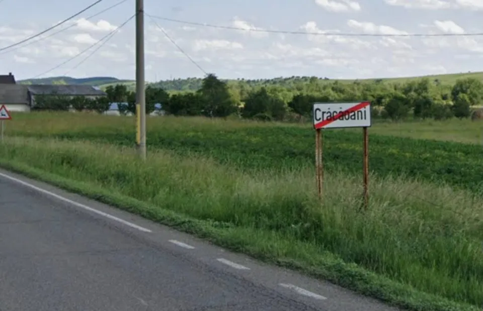 Un semn de intrare într-o localitate cu textul Crăcăoani este situat pe marginea unui drum asfaltat lângă un câmp de iarbă verde.