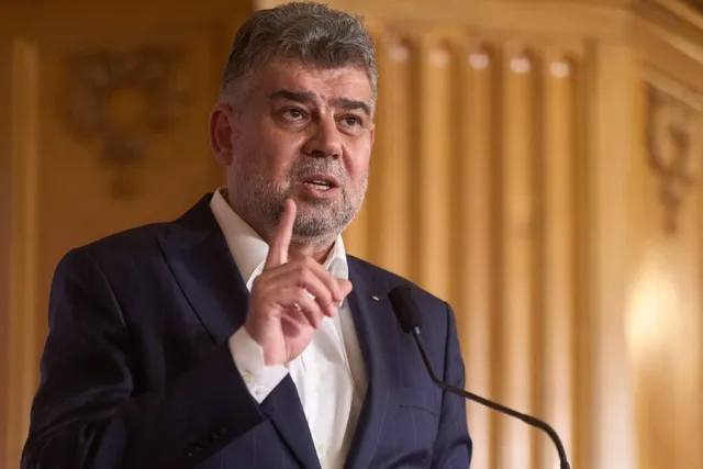 Un bărbat cu părul scurt și barbă stufoasă poartă un sacou închis și cămașă albă, ridicând un deget în fața unui microfon.
