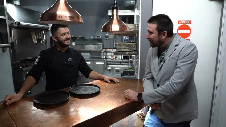 Un bărbat în halat negru de bucătar stă în spatele unui blat de cupru, în timp ce un alt bărbat într-un sacou gri stă în fața acestuia.