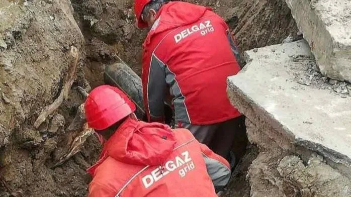 Două persoane purtând veste de lucru roșii cu inscripții albe sunt așezate într-o șanț excavat în pământ.