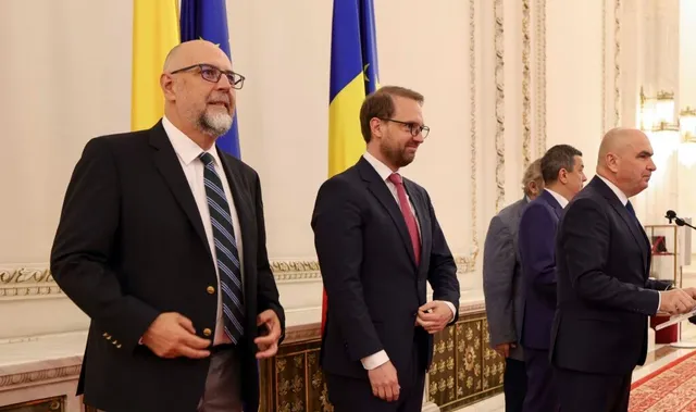Patru bărbați în costume oficiale stau în picioare într-o încăpere cu panouri decorative și steaguri colorate în fundal.