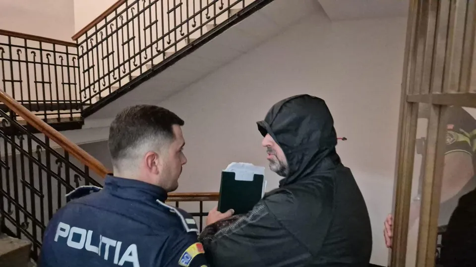 Un agent de poliție în uniformă albastră stă față în față cu un bărbat îmbrăcat într-o hanoracă neagră lângă o scară interioară cu balustradă metalică.