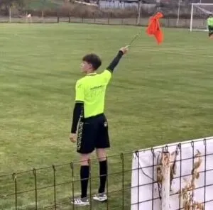 Un bărbat îmbrăcat în tricou galben și pantaloni scurți negri ridică un obiect lung și subțire spre cer pe un teren de fotbal.