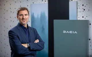 Un bărbat cu sacou albastru și cămașă închisă stă cu brațele încrucișate în fața unui panou gri cu logo-ul Dacia.