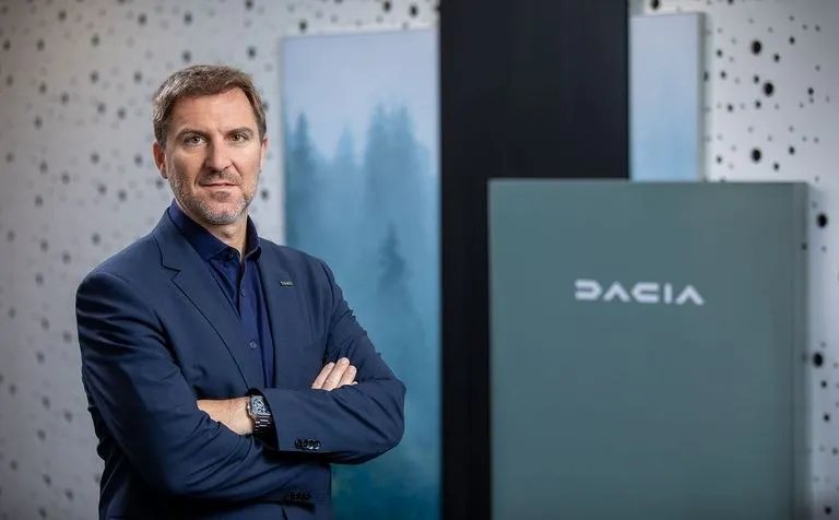 Un bărbat cu sacou albastru și cămașă închisă stă cu brațele încrucișate în fața unui panou gri cu logo-ul Dacia.