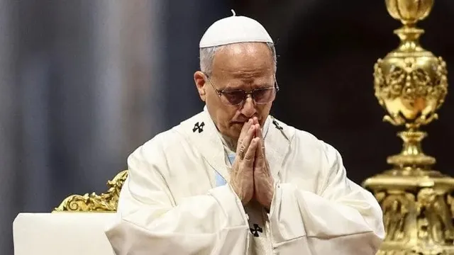 Un bărbat cu ochelari și o căciulă albă poartă haine albe și stă cu mâinile împreunate sub bărbie într-o poziție de rugăciune.