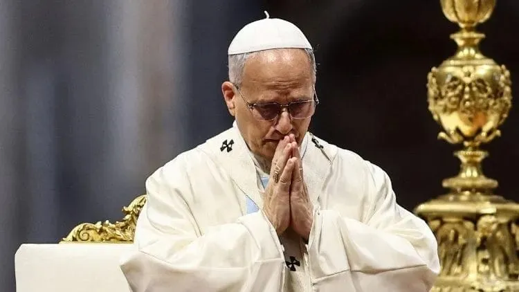 Un bărbat cu ochelari și o căciulă albă poartă haine albe și stă cu mâinile împreunate sub bărbie într-o poziție de rugăciune.