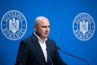 Un bărbat cu părul scurt și pielea deschisă, purtând sacou închis și cămașă albă, stă în fața unui fundal albastru cu două embleme circulare.