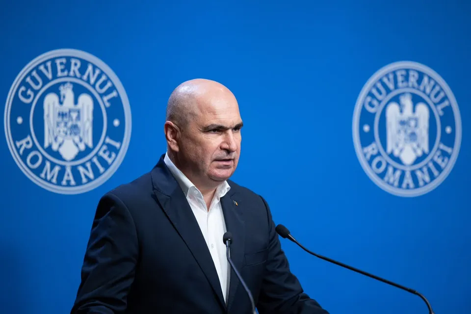 Un bărbat cu părul scurt și pielea deschisă, purtând sacou închis și cămașă albă, stă în fața unui fundal albastru cu două embleme circulare.