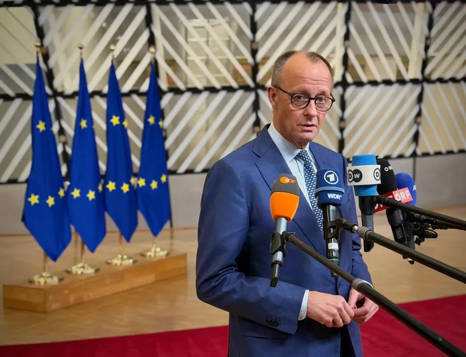 Un bărbat cu ochelari și costum albastru stă în fața mai multor microfoane și a unor steaguri albastre cu stele galbene.