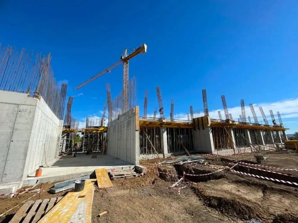 Un șantier cu structuri de beton gri, stâlpi de armătură metalică și o macararie galbenă sub un cer albastru.