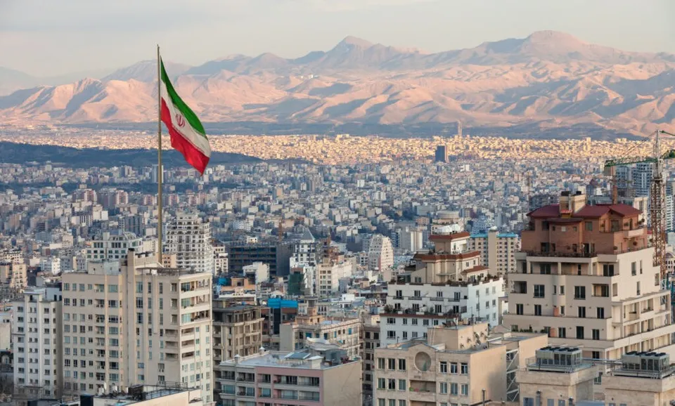 O steag iranianagândește pe un stâlp înalt deasupra unui peisaj urban dens cu clădiri și munți în fundal.