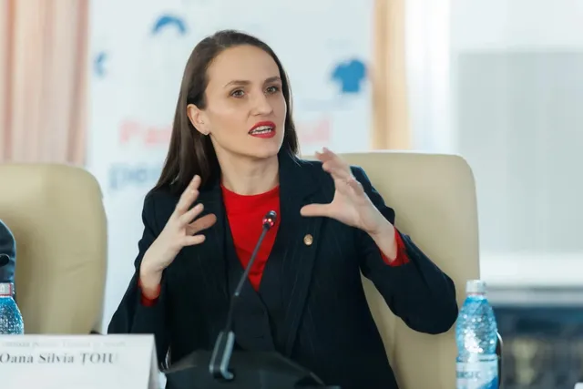 O femeie cu părul lung și o jachetă neagră peste un tricou roșu stă la o masă cu un microfon și o sticlă de apă.