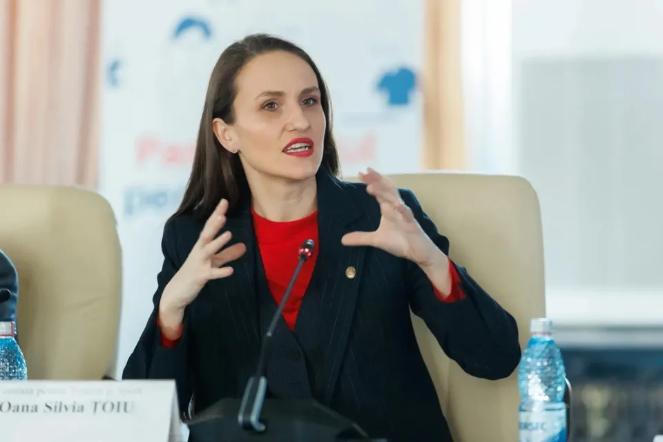 O femeie cu părul lung și o jachetă neagră peste un tricou roșu stă la o masă cu un microfon și o sticlă de apă.