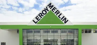 Fațada unei clădiri albe cu un triunghi verde pe care este scris textul negru LEROY MERLIN sub un cer cu nori.
