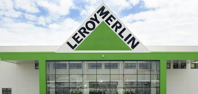 Fațada unei clădiri albe cu un triunghi verde pe care este scris textul negru LEROY MERLIN sub un cer cu nori.