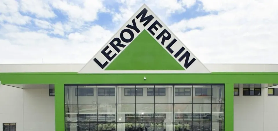 Fațada unei clădiri albe cu un triunghi verde pe care este scris textul negru LEROY MERLIN sub un cer cu nori.