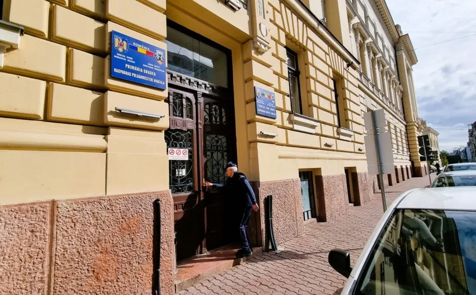 Un bărbat cu căciulă și jachetă închisă pășește prin ușa de lemn a unei clădiri galbene cu plăcuțe informative pe perete.