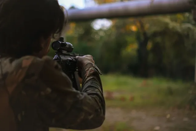 O persoană îmbrăcată în haine de camuflaj țintește cu o armă echipată cu lunetă spre un peisaj de pădure desfășurat în fundal.