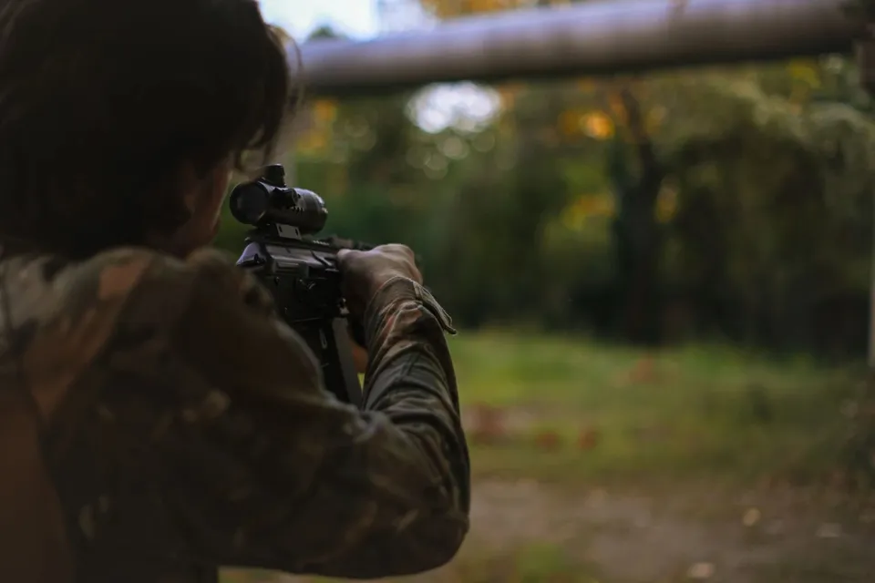 O persoană îmbrăcată în haine de camuflaj țintește cu o armă echipată cu lunetă spre un peisaj de pădure desfășurat în fundal.
