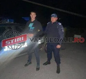 Un bărbat cu hanorac negru și un agent de poliție în uniformă albastră stau lângă o mașină în timpul nopții.