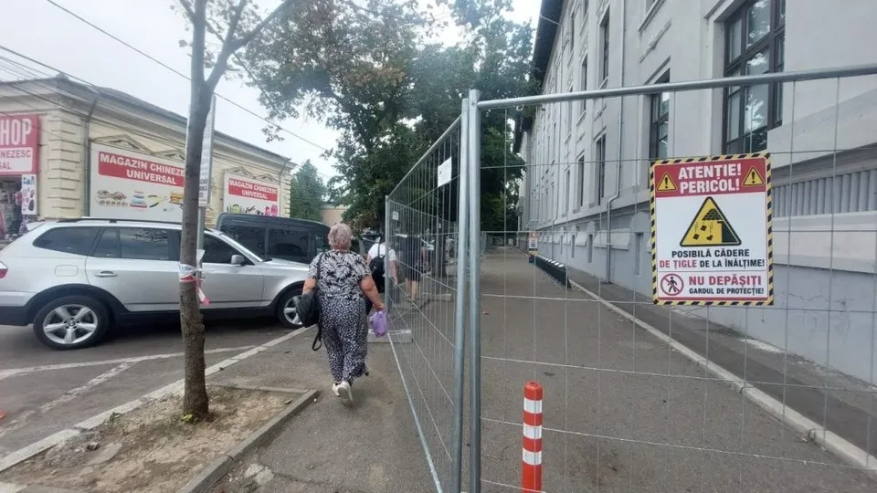 O femeie merge pe un trotuar lângă un gard metalic de șantier, în timp ce mașini sunt parcate pe partea opusă a drumului.