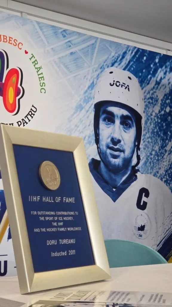 Un cadru cu un trofeu albastru și un medaliu pe care scrie IIHF Hall of Fame și Doru Tureanu este așezat în fața unui poster.