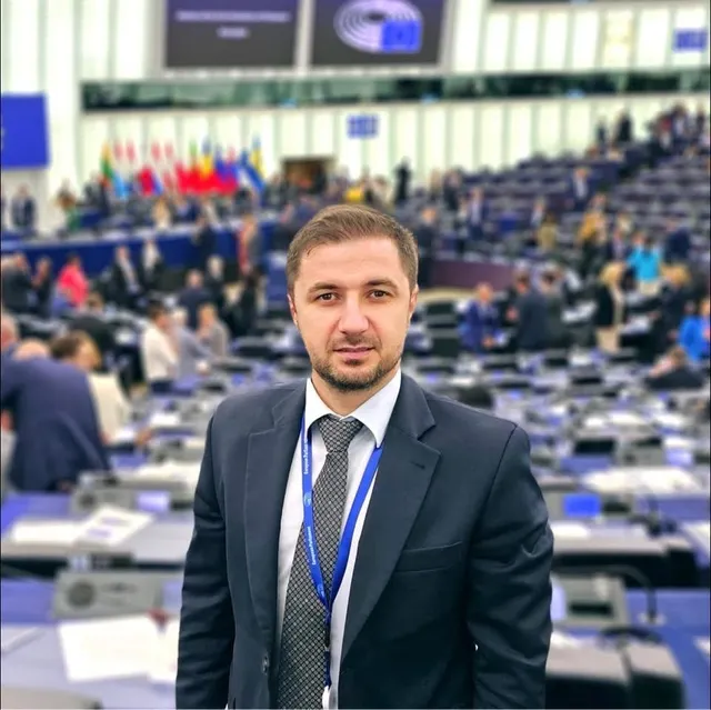 Un bărbat cu barbă, purtând un costum închis la culoare, cămașă albă, cravată și un cordon albastru la gât, stă în fața unei săli de conferințe ocupate.