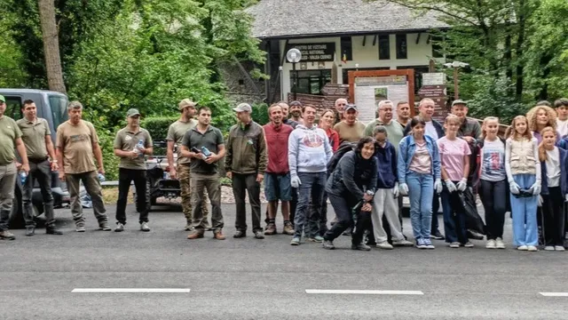 Un grup de oameni stă în rând pe un drum asfaltat în fața unei clădiri de lemn înconjurate de copaci verzi.