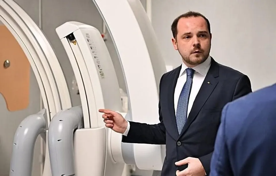 Un bărbat într-un costum închis și cravată albastră arată spre un aparat medical alb de dimensiuni mari.