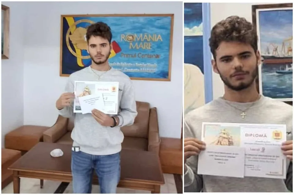 Un bărbat cu barbă poartă un hanorac gri și pantaloni albaștri, ținând două diplome albe în mâini în fața unui tablou și al unei canapele maro.