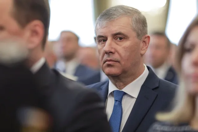 Un bărbat cu părul scurt și gri, purtând sacou închis la culoare și cravată albastră, privește spre camera de filmare într-un decor interior.