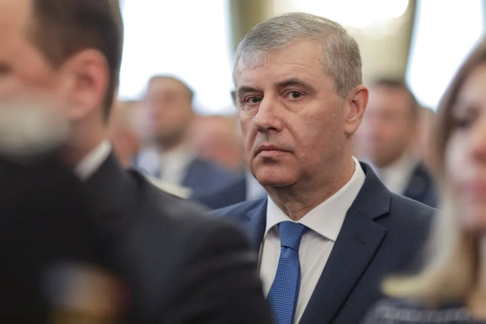 Un bărbat cu părul scurt și gri, purtând sacou închis la culoare și cravată albastră, privește spre camera de filmare într-un decor interior.