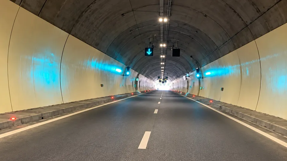 O porțiune de drum asfaltat cu linii albe marcate traversează un tunel cu pereți curbați și lumină artificială suspendată pe tavan.
