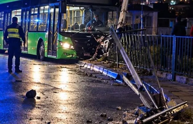 Un autobuz verde este oprit pe un drum umed lângă un stâlp de utilități înclinat și o gardă metalică dărâmată noaptea.