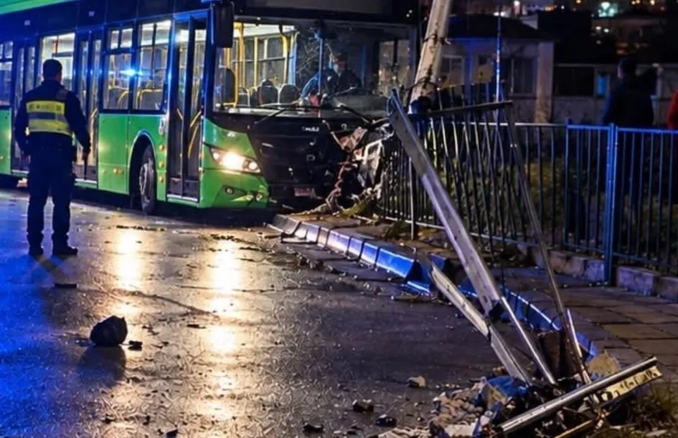 Un autobuz verde este oprit pe un drum umed lângă un stâlp de utilități înclinat și o gardă metalică dărâmată noaptea.