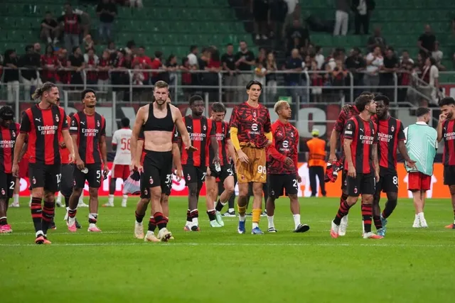 Un grup de fotbaliști în echipament roșu și negru stau pe terenul de fotbal sub luminile unui stadion plin de spectatori.
