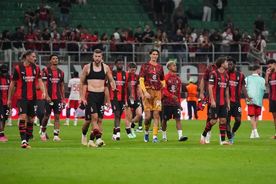 Un grup de fotbaliști în echipament roșu și negru stau pe terenul de fotbal sub luminile unui stadion plin de spectatori.