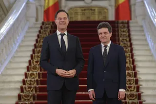 Doi bărbați în costume închise stau în picioare în fața unei scări interioare cu covor roșu și balustrade albe.