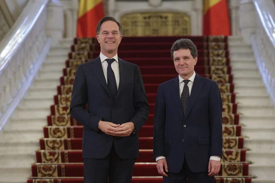 Doi bărbați în costume închise stau în picioare în fața unei scări interioare cu covor roșu și balustrade albe.