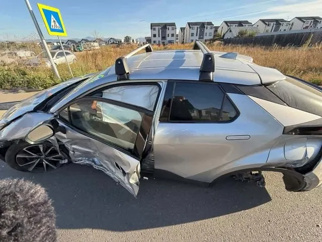 O mașină argintie cu partea din față și portierele stângi zdrobite stă pe un drum asfaltat lângă vegetație și clădiri rezidențiale.