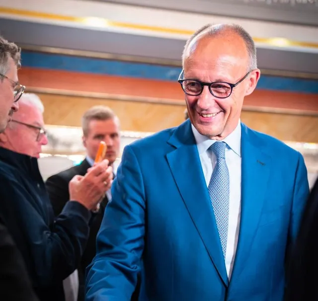 Un bărbat zâmbind cu ochelari și costum albastru este încadrat de alți bărbați într-un spațiu interior iluminat.