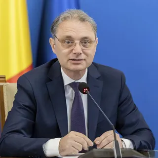Un bărbat în costum bleumarin, cravată violet și ochelari, stă la o masă cu un microfon, cu steagul României în fundal.
