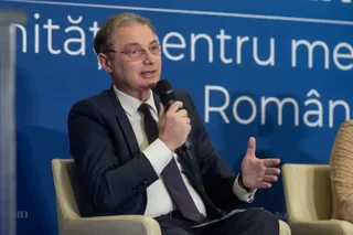 Un bărbat cu ochelari, purtând costum închis la culoare și cravată, vorbește într-un microfon stând așezat într-un fotoliu de culoare deschisă.