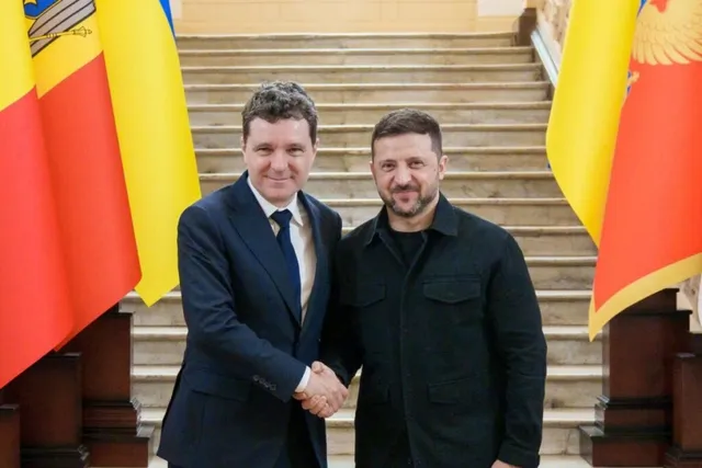 Un bărbat într-un costum albastru și unul într-o cămașă neagră stau pe trepte și își strâng mâinile.
