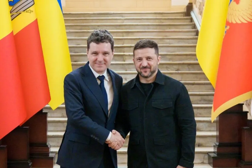 Un bărbat într-un costum albastru și unul într-o cămașă neagră stau pe trepte și își strâng mâinile.
